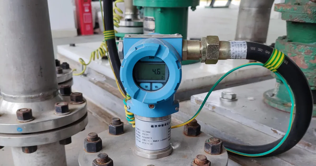 Industrial field installation of a magnetostrictive level gauge mounted on a pipe flange, featuring a localized digital LCD indicator and intrinsically safe wiring.