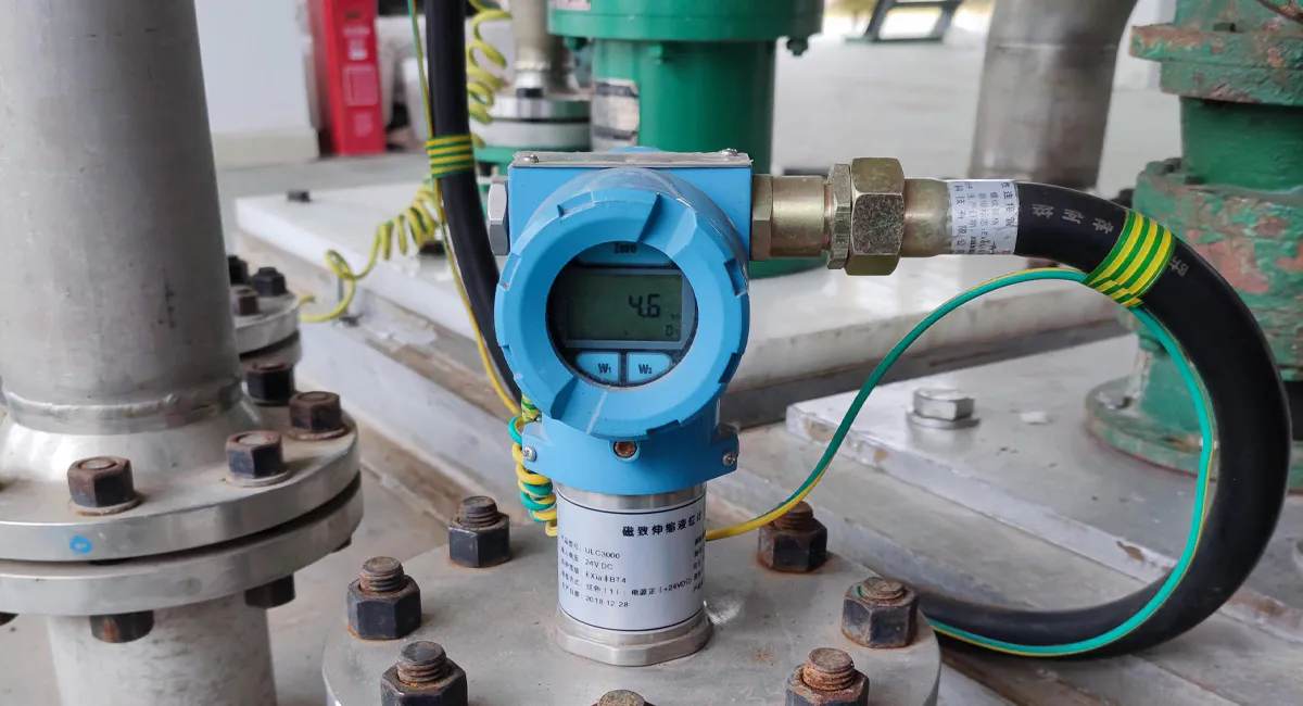 Industrial field installation of a magnetostrictive level gauge mounted on a pipe flange, featuring a localized digital LCD indicator and intrinsically safe wiring.